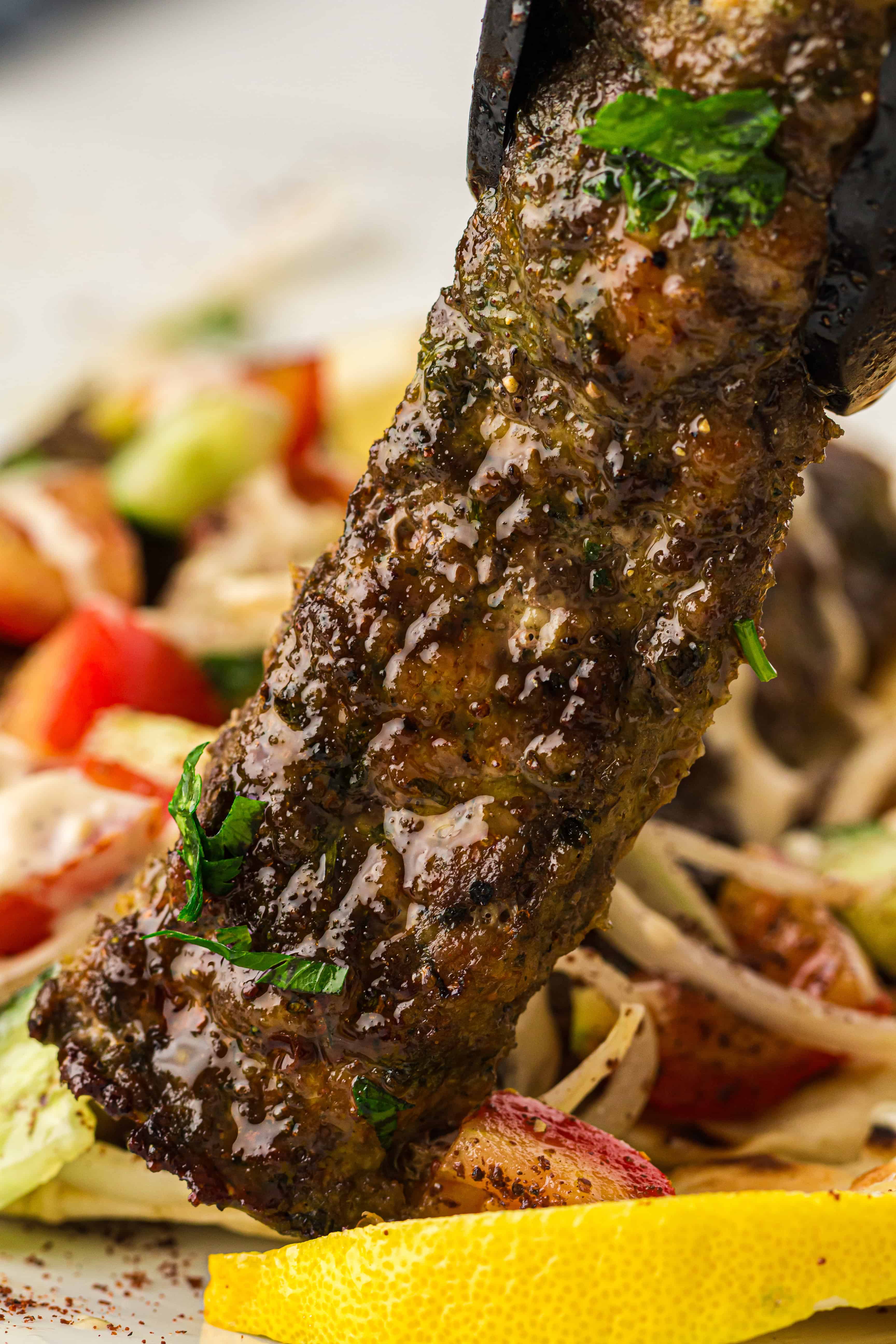 A close-up of a grilled, seasoned meat skewer garnished with herbs, held above a plate of salad and lemon wedges.