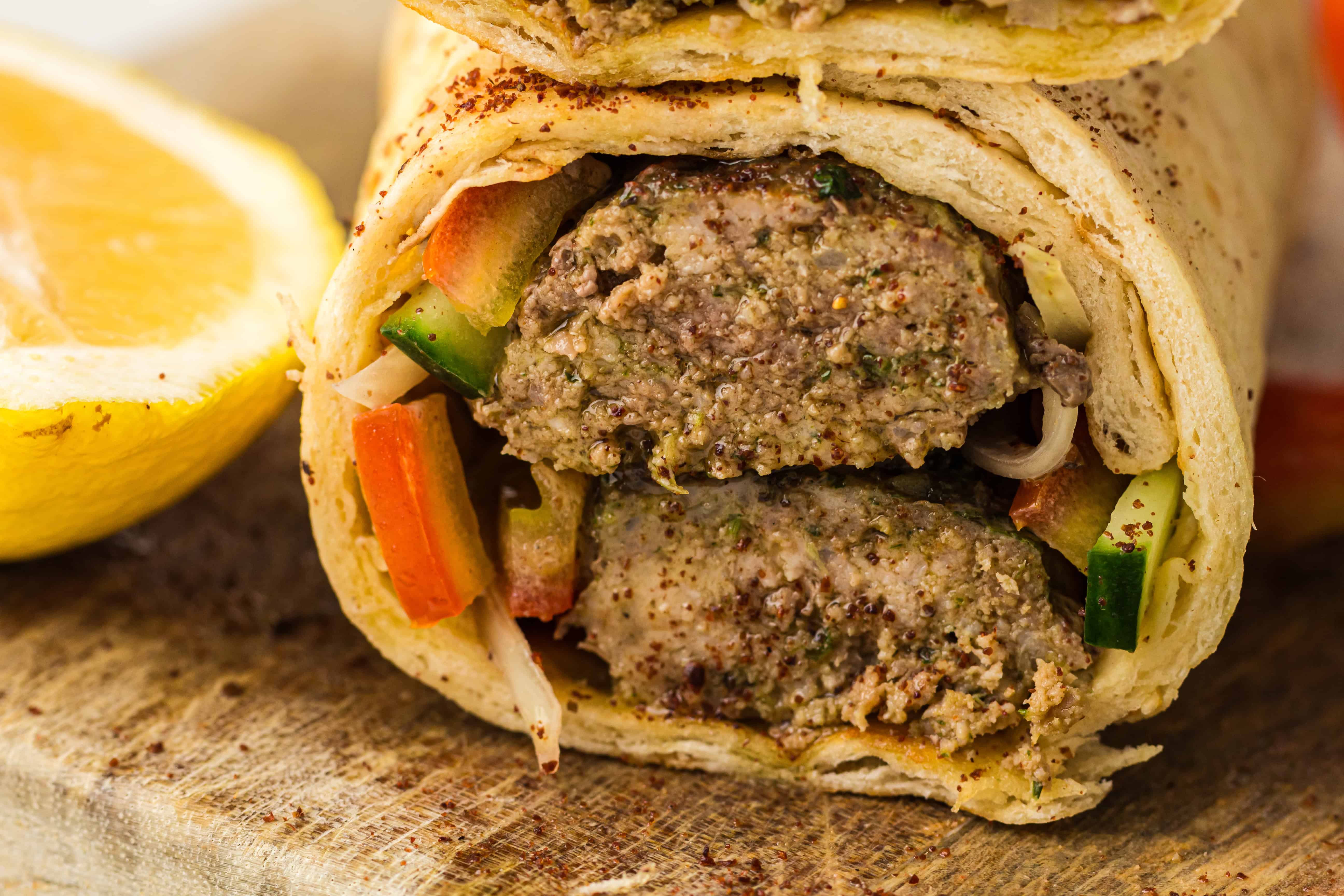 Close-up of a kebab wrap with ground meat, sliced cucumbers, tomatoes, and a lemon wedge on the side.