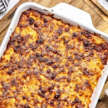 Baked macaroni and cheese in a white dish on a wooden board, with parsley and uncooked pasta nearby.