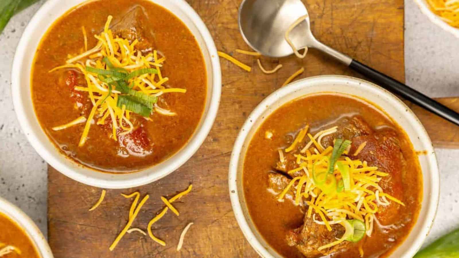 Bowls of chili topped with shredded cheese and green onions, with a spoon on a wooden board.
