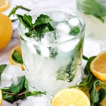 A glass of lemonade with ice, mint leaves, lemon slices, and fresh mint on a marble surface.