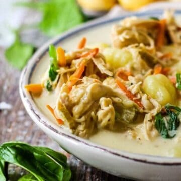 Creamy soup with shredded chicken, gnocchi, spinach, and carrots in a white bowl on a rustic wooden table.