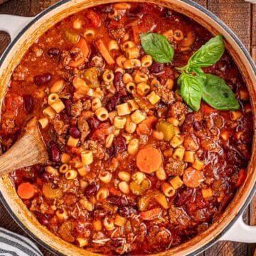 A pot of pasta soup with vegetables, beans, and ground meat, garnished with fresh basil leaves.