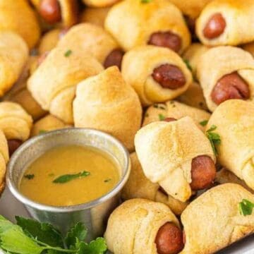 Mini pigs in a blanket served on a white platter with a cup of mustard dipping sauce and fresh parsley garnish.