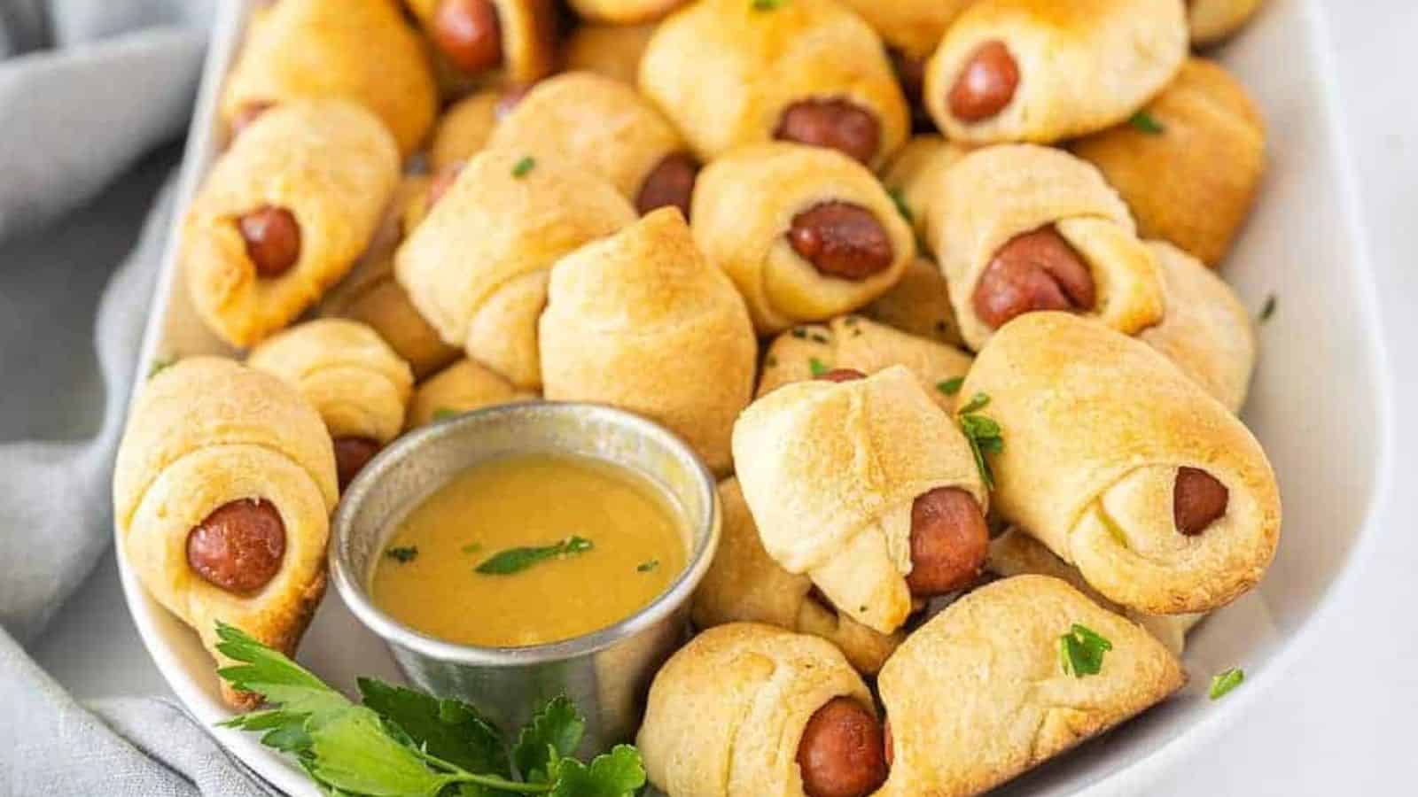 Mini pigs in a blanket served on a white platter with a cup of mustard dipping sauce and fresh parsley garnish.