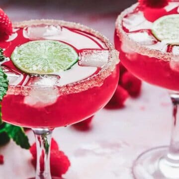 Two glasses of pink cocktail with ice, lime slices, sugared rims, mint, and raspberries as garnish.