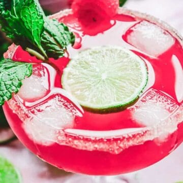 A bright pink cocktail with ice, a lime slice, mint leaves, and a sugared rim.