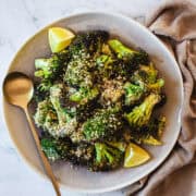 A plate of roasted broccoli topped with sesame seeds and lemon wedges, with a gold spoon on the side.