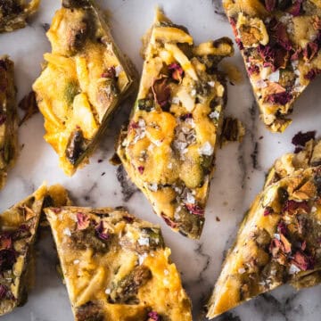 Close-up of sliced dessert bars resembling pistachio rose bark, topped with sea salt and dried flower petals on a marble surface.