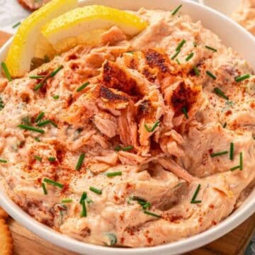 Creamy salmon dip garnished with chives and lemon slices, served with round crackers on the side.