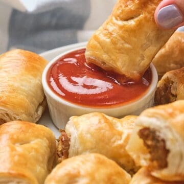 A hand dips a golden sausage roll into a bowl of red sauce, surrounded by more sausage rolls on a plate.