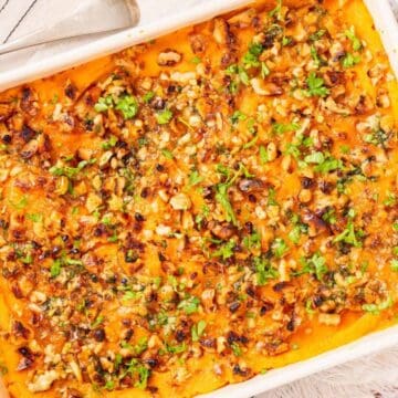 Baked sweet potato casserole topped with chopped nuts and herbs in a white dish, with a serving spoon beside it.
