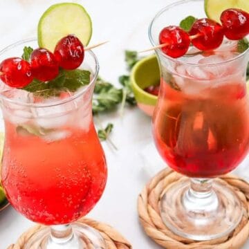 Two red cocktails with ice, garnished with lime, cherries, and mint, on woven coasters with lime slices nearby.
