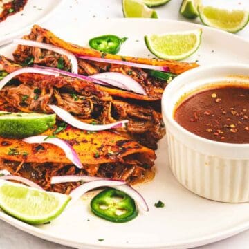 Three birria tacos with lime wedges, sliced onions, and dipping sauce on a white plate.