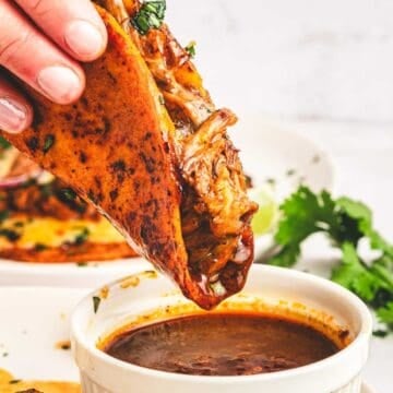 Hand dipping a birria taco filled with meat into a bowl of consommé, with limes and cilantro in the background.
