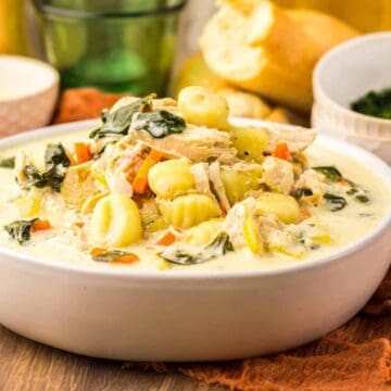 Creamy chicken gnocchi soup with vegetables served in a white bowl, bread and herbs on the side.