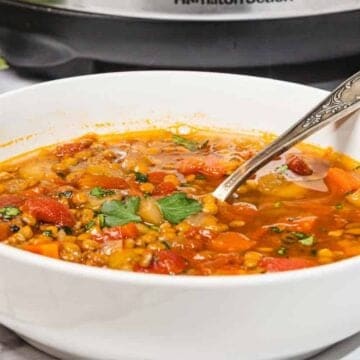 A bowl of vegetable lentil soup with a spoon, garnished with herbs, on a marble countertop.