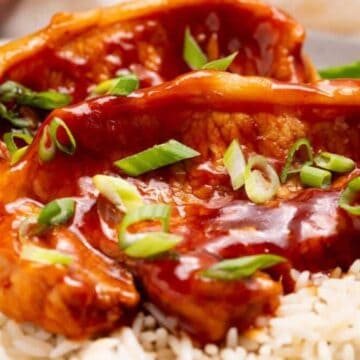 Two pork chops with reddish sauce and sliced green onions served over white rice on a plate.