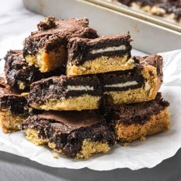 A stack of Slutty Brownies with visible sandwich cookies in the middle, layered between fudgy chocolate chip cookie brownies, sits on parchment paper.