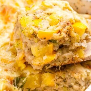 A wooden spoon lifts a cheesy, baked squash casserole from a white speckled baking dish.