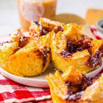 Close-up of flaky pastries with jam filling on a white plate, set on a red checked cloth with powdered sugar nearby.
