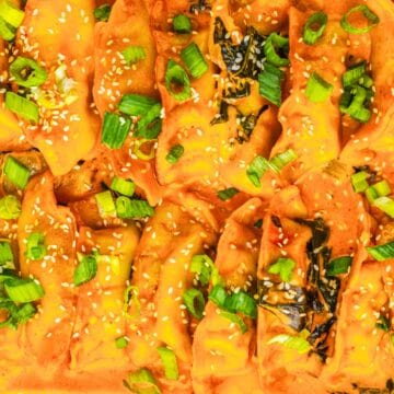 Close-up of baked dumplings in orange sauce, topped with chopped green onions and sesame seeds.