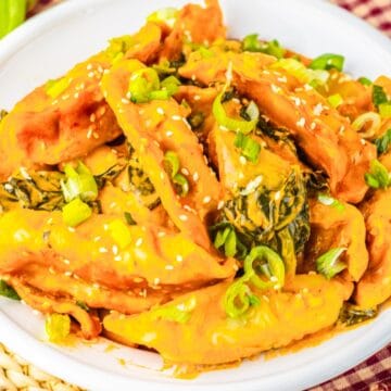 A white plate of dumplings in orange sauce, topped with chopped green onions and sesame seeds.