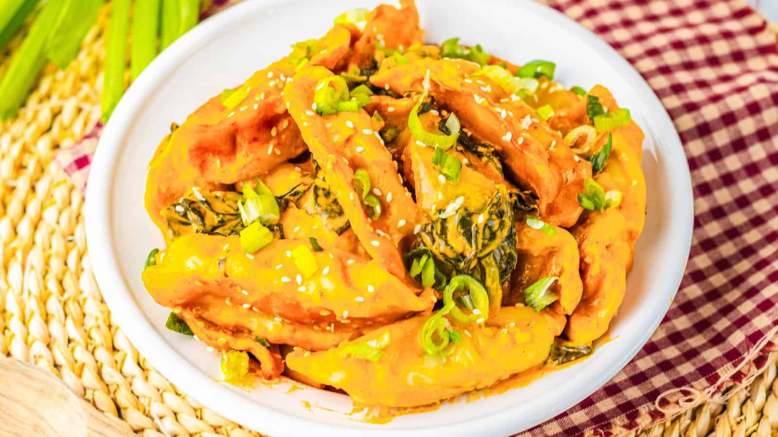 A white plate of dumplings in orange sauce, topped with chopped green onions and sesame seeds.