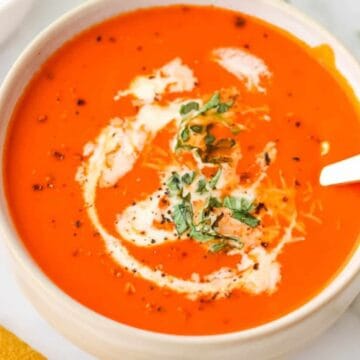 A bowl of creamy tomato soup garnished with herbs and pepper, with a spoon resting inside.