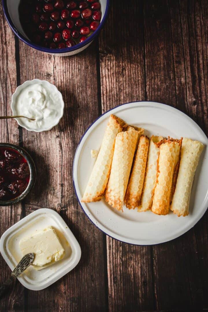 Five traditional Russian rolled wafers sit on a plate, surrounded by bowls of cherries, cream, jam, and a dish of butter atop a rustic wooden table.