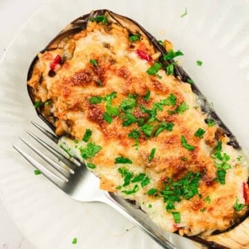 A baked stuffed eggplant topped with melted cheese and chopped herbs, served on a white plate with a fork.