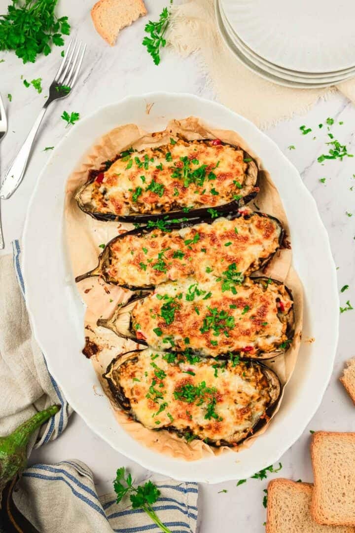 Four baked stuffed eggplant halves topped with melted cheese and chopped parsley on a white oval dish.