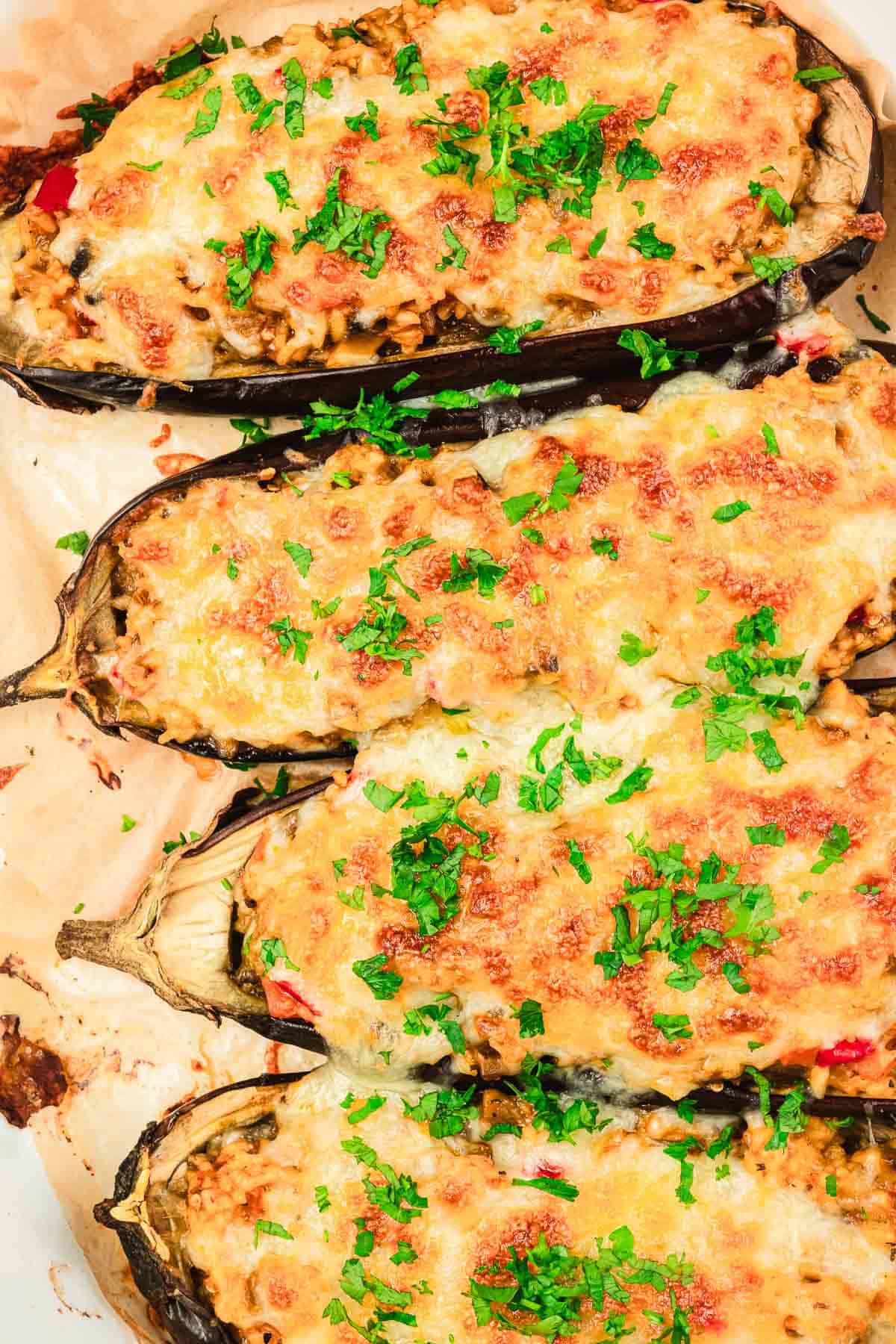 Baked stuffed eggplants topped with melted cheese and chopped fresh parsley on parchment paper.