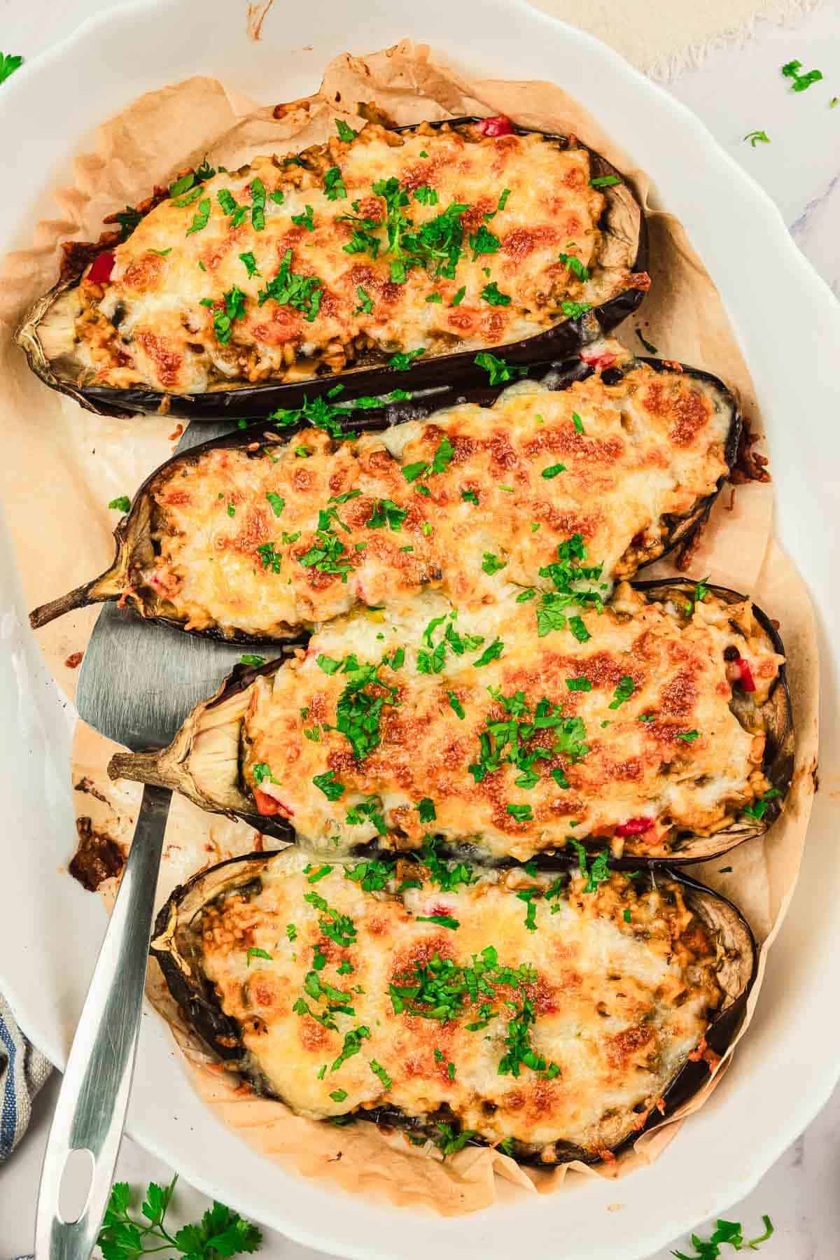 Four baked stuffed eggplants topped with melted cheese and chopped herbs, served on a white oval dish.
