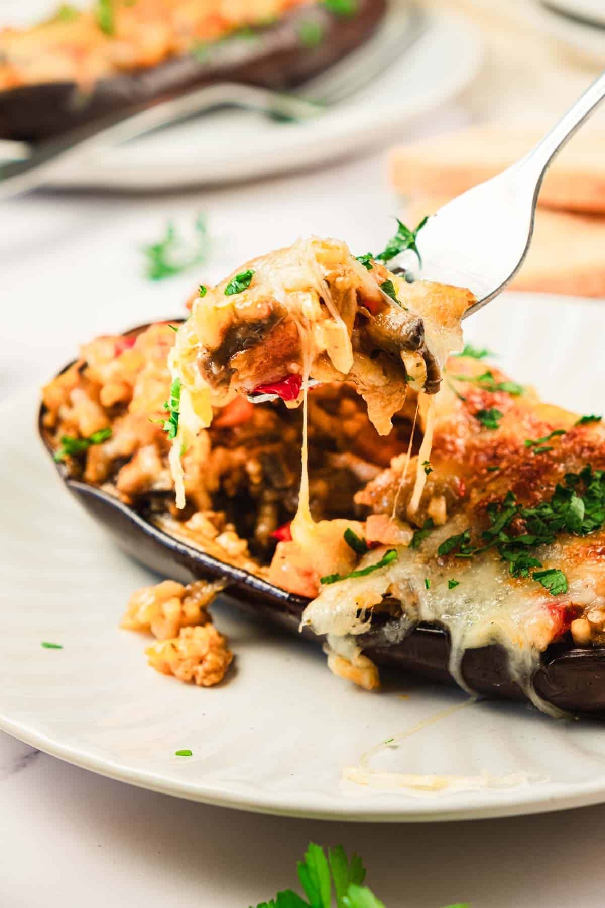 Stuffed eggplant with melted cheese, rice, and vegetables on a plate; a fork holds a cheesy bite.