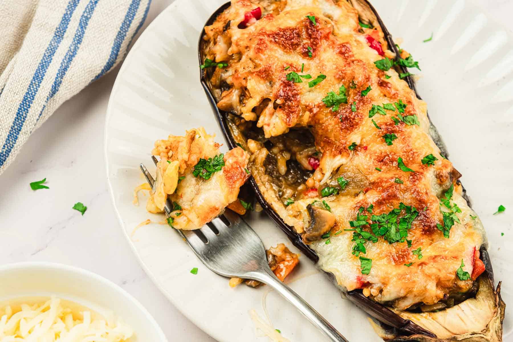 A plate with a stuffed, baked eggplant topped with melted cheese and parsley, next to a forkful of the dish.