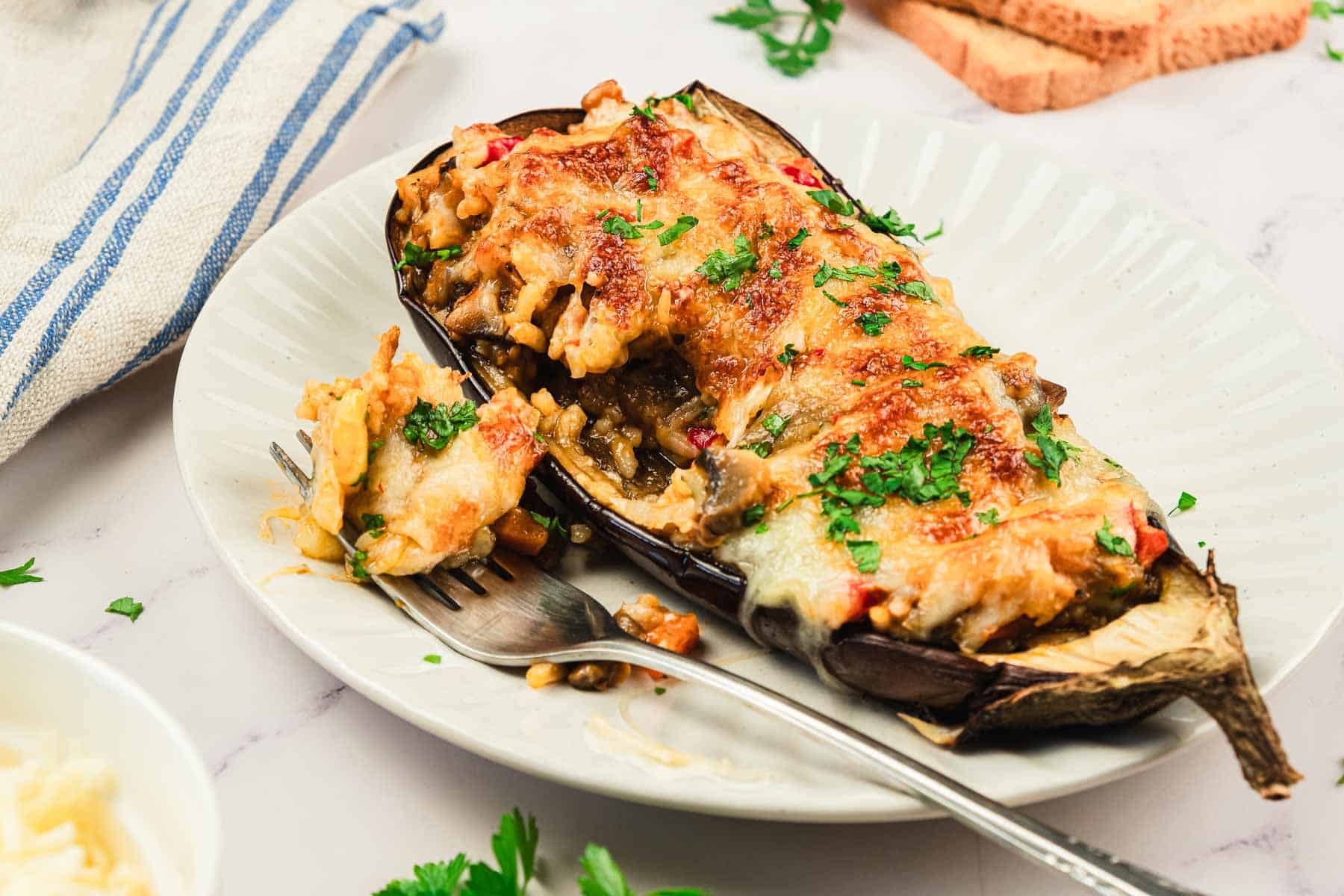 Baked stuffed eggplant topped with melted cheese and herbs on a plate with a fork.