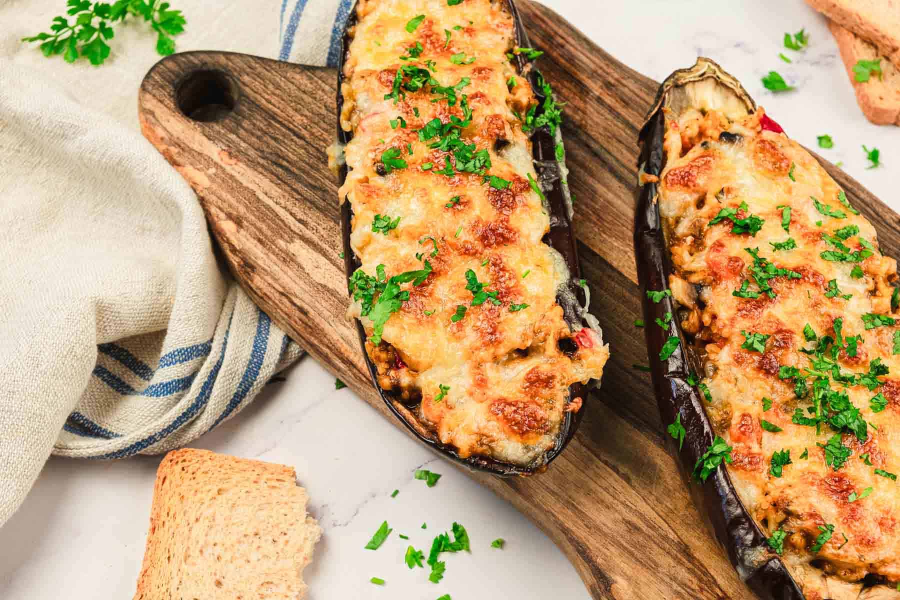 Two baked stuffed eggplants topped with melted cheese and chopped parsley on wooden boards.