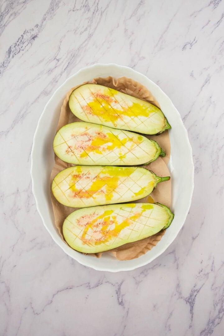 Four halved eggplants with scored flesh, drizzled with oil in a white oval baking dish on a marble surface.