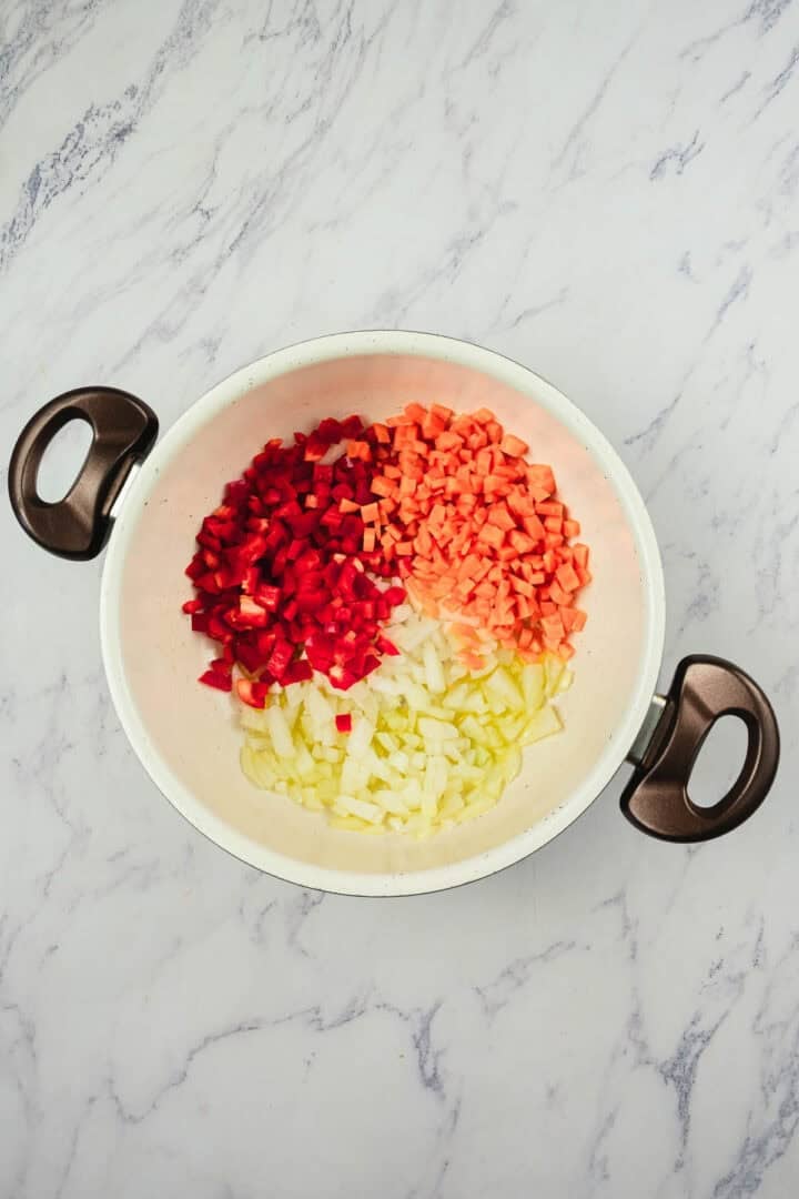 White pan with chopped onions, carrots, and red bell peppers, placed on a marble countertop.