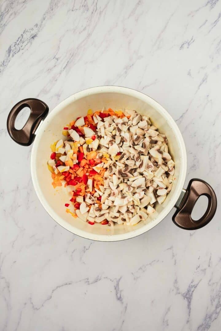 A white pot with chopped mushrooms, onions, and red bell peppers on a marble countertop.