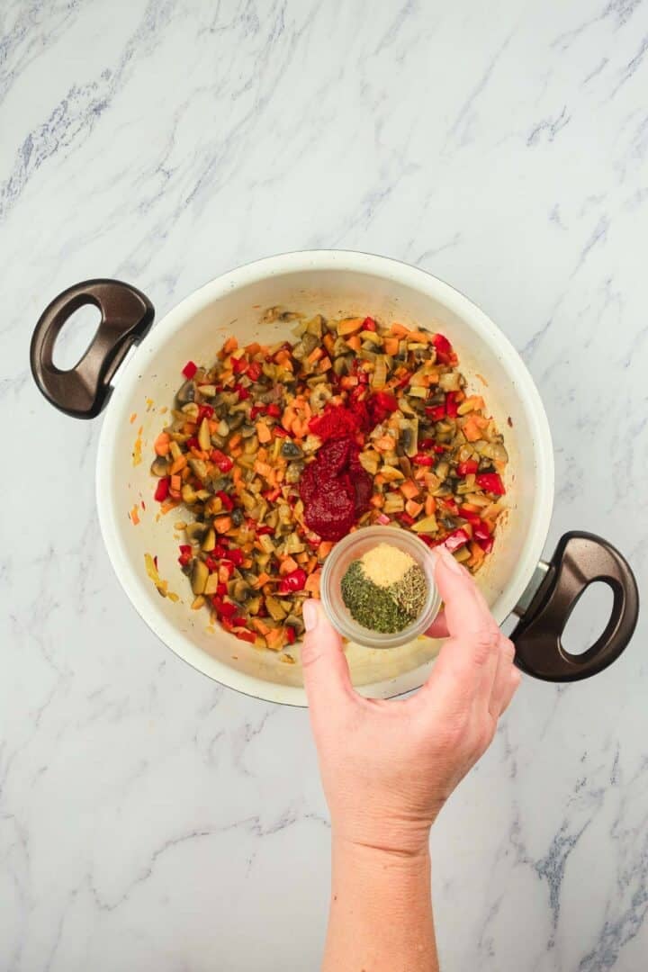 A hand adds dried herbs to a pot of chopped vegetables and tomato paste on a marble countertop.