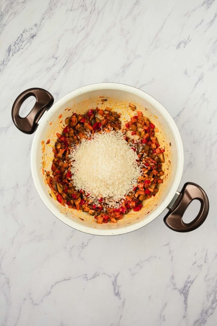 A pot with chopped vegetables and a mound of uncooked white rice on a marble countertop.