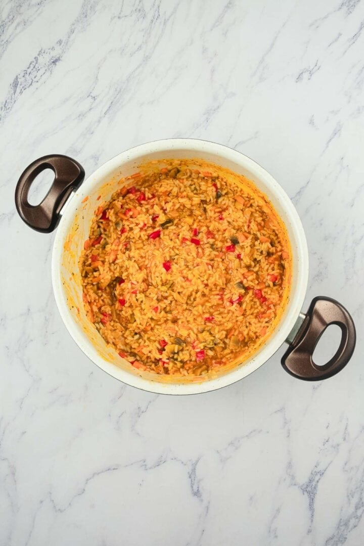 A pot of cooked rice mixed with red and green peppers on a white marble surface.