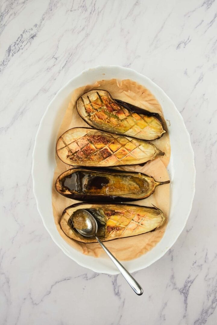 Three roasted eggplant halves with scored tops on parchment in a white oval dish, with a spoon beside them.