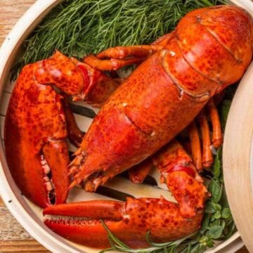 Steamed whole lobster in a bamboo steamer basket with fresh herbs on a wooden table.
