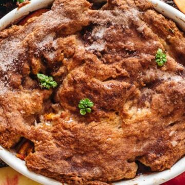 An overhead view of a freshly baked peach cobbler in a round baking dish. The cobbler's surface is golden-brown with a sprinkling of sugar, and it is garnished with a few small green herb leaves. Whole and sliced peaches surround the dish, reminiscent of Forgotten Dessert Classics.