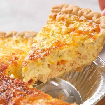 A hand lifts a slice of cheesy, golden-brown quiche from a pie tin.