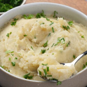 A bowl of creamy mashed potatoes topped with chopped parsley, with a spoonful being lifted.