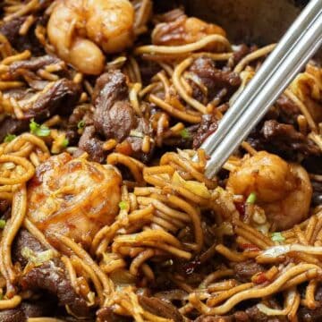 Close-up of stir-fried noodles with shrimp, beef, and vegetables, mixed with chopsticks in a pan.